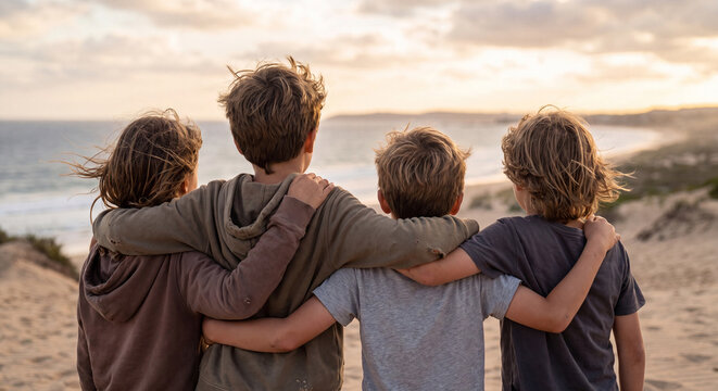 Three young brothers or friends sit side by side on a sandy beach looking towards the sea during sunset