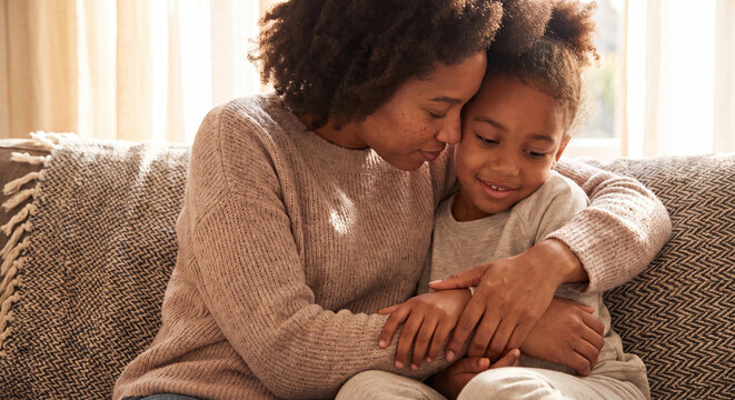 A loving mother gently hugging and kissing her young daughter while sitting together on a comfortable sofa at home