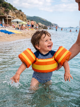 A happy young toddler wearing striped orange inflatable armbands splashes joyfully in the ocean water during a summer beach vacation.