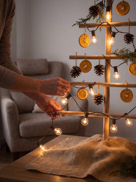 Close up of hands wrapping warm string lights around a handcrafted wooden dowel Christmas tree adorned with dried orange slices and pine cones