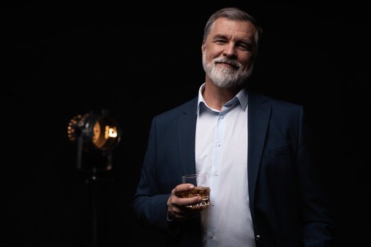 Handsome businessman in stylish suit holding glass whiskey on dark