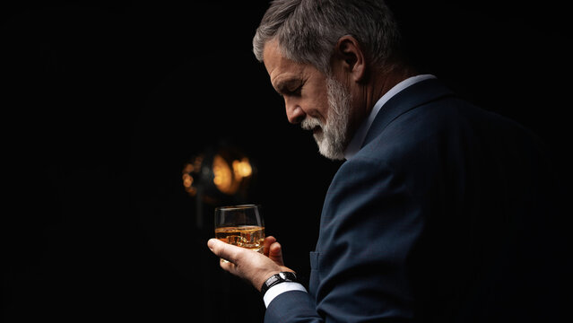 Handsome businessman in stylish suit holding glass whiskey on dark