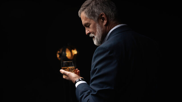 Handsome businessman in stylish suit holding glass whiskey on dark