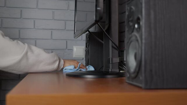 Person cleaning a dusty wooden computer desk with a microfiber cloth. Person's hand cleaning a brown wooden office desk with a blue microfiber cloth, wiping dust around a computer monitor and speakers