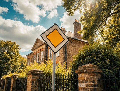 Yellow diamond priority road sign stands in front of old brick building. Green trees, bushes surround historical architecture under bright cloudy sky. Summer sun shines through leaves near street