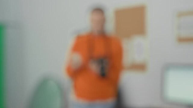 Man in orange sweater holding dslr with left hand and tapping smartphone with right hand amid green screen and reflector in studio; concentration.