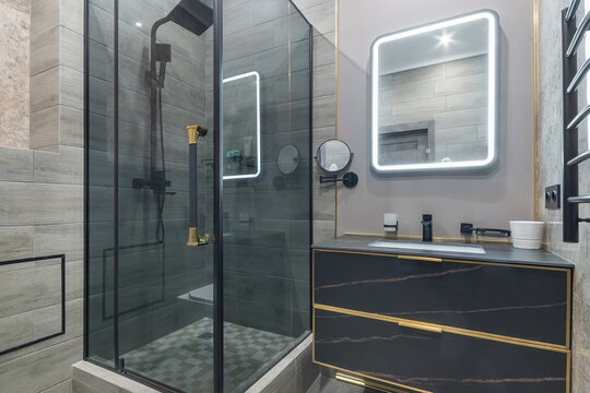 A bathroom features a glass shower with black fixtures. A dark vanity with gold accents is next to a well-lit mirror. The space is clean and shows a modern style