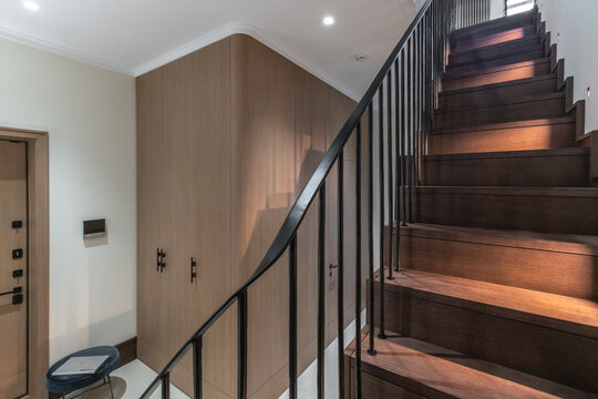A staircase with wooden steps leads to an upper area in a modern home. The hallway is well-lit and features a built-in storage unit. The space has a clean layout