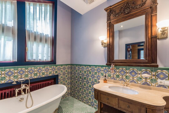 A vintage bathroom features a freestanding bathtub and a wooden sink area. The walls and tiles show colorful patterns. Natural light comes in through the windows