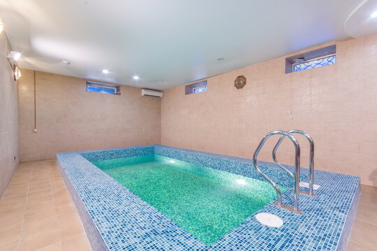 A small indoor pool is in a room with light-colored walls. The water is clear and inviting. There are steps leading into the pool. The room has windows for natural light