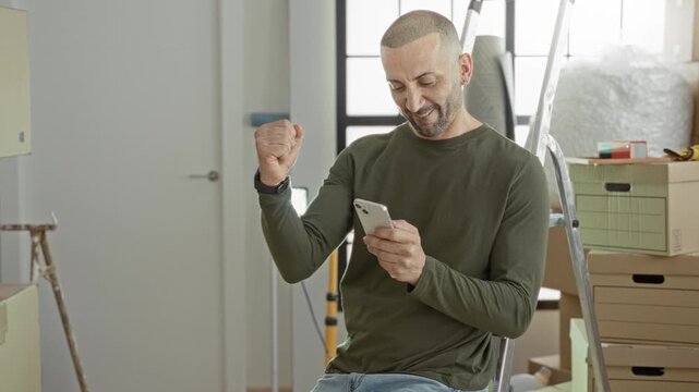 Man holding smartphone, fist pump gesture while seated amid cardboard boxes and ladder in building; new start joy.