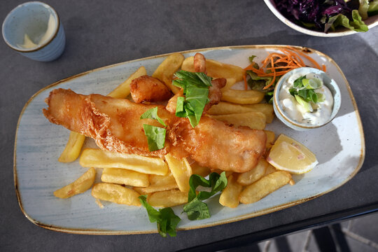 Fish and chips, portion of fried cod fillet and handmade French fries, served with remoulade and a lemon wedge on a gray platter, top view from above