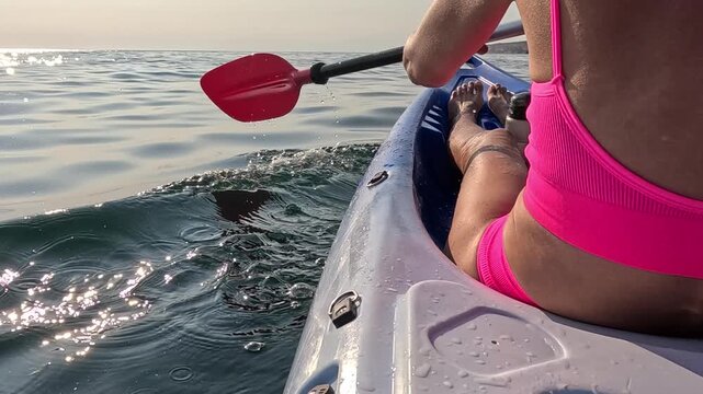 Pink bikini athlete propels blue kayak across shimmering Mediterranean sea surface at dawn. Close-up over-the-shoulder point of view shot.