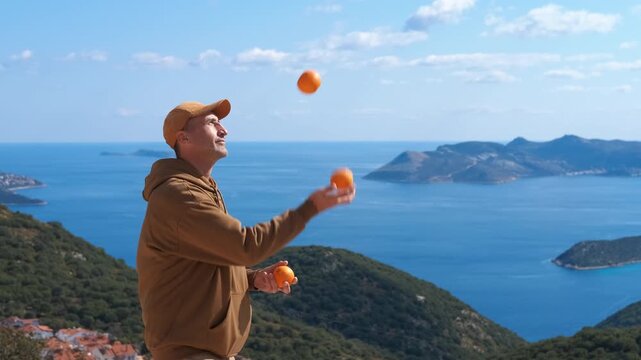 Man juggling oranges with beautiful sea view. Young man in casual clothes juggling with three oranges, standing on a hill with a beautiful sea view