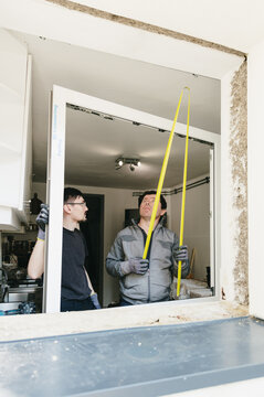 Vertical portrait of two workers measuring a window frame with a yellow tape measure.
