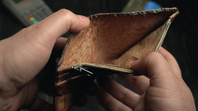 Close-up of hands opening a brown leather wallet, revealing the interior and zipper compartment, with credit cards and a payment terminal visible in the background on a dark wooden surface