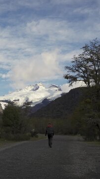 Back view of a hiker with a backpack walking along a dirt trail towards the snow-capped peak of Cerro Tronador, surrounded by forest in Patagonia, Argentina.