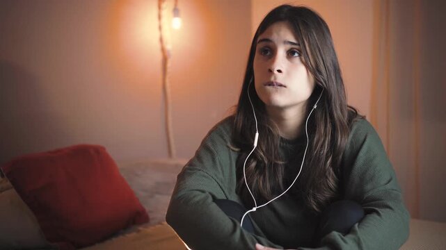 Young Hispanic woman in her bedroom crying while listening to music with headphones. Emotional moment, sadness, self-reflection, mental health and personal feelings.
