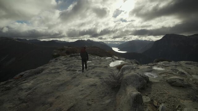 Back view of a hiker standing on a high rocky cliff at La Mirada del Doctor viewpoint, observing the expansive mountain range and lake in Nahuel Huapi National Park, Argentina.