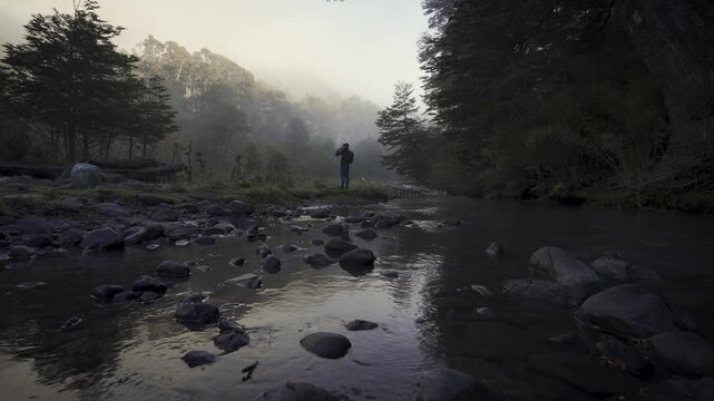 Hiker with a backpack stands on the rocky bank of a river in a misty autumn forest, surrounded by tall trees and natural wilderness.