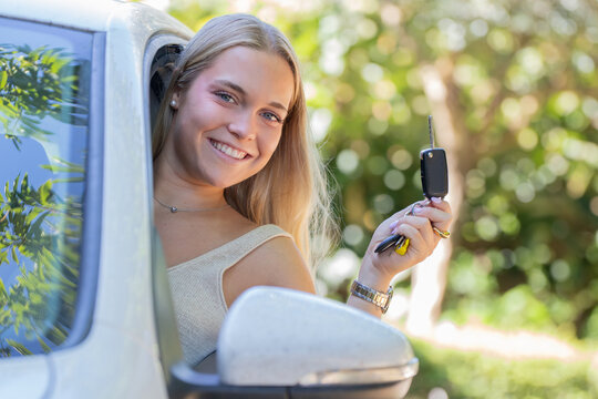 Happy young woman smiling and leaning out of a car window while holding up car keys, celebrating getting her first car or driver's license