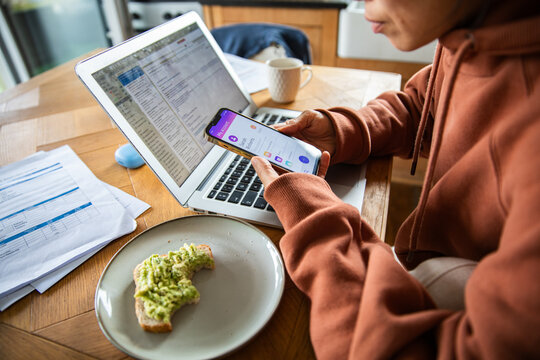 Person managing finances on smartphone and laptop at home kitchen table