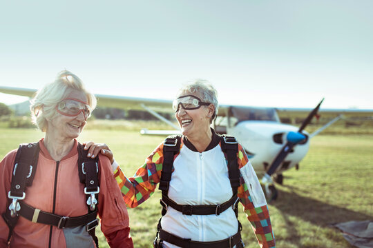 Two senior women in skydiving gear at rural airfield