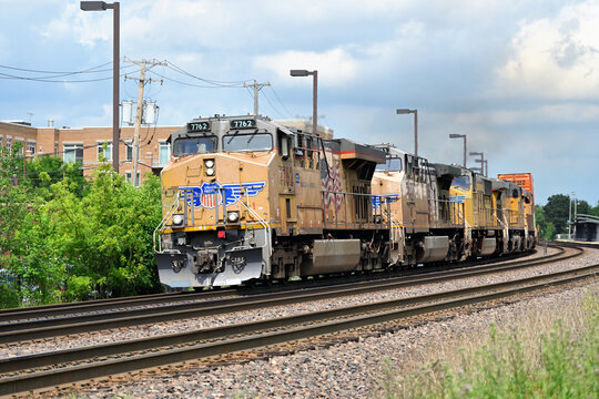 A quintet of Union Pacific Railroad locomotive units lead a westbound container train past a local commuter rail station in the western Chicago suburban area.