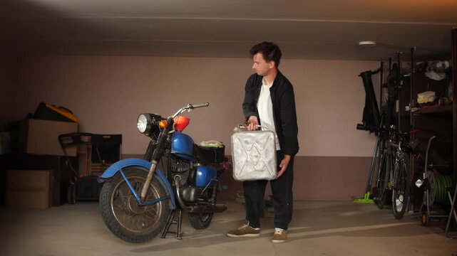 Young man standing in a garage, pouring fuel from a metal gas can into the tank of a blue vintage motorcycle, performing maintenance or restoration on the classic vehicle