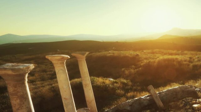 Two stone columns stand in a grassy field as the sun sets behind distant hills. The sky shows warm colors, marking the end of the day with a blend of natural beauty and history.
