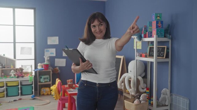 Woman with clipboard points finger toward toy shelf by blue wall in kindergarten building while holding a pen; warmth guidance.