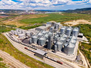 Industrial facility with silos and railway tracks near a green landscape in a rural area under a blue sky with clouds © DSMT