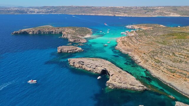 Blue Lagoon, Comino, Malta, with turquoise Mediterranean water, boats and rocky islets near Cominotto, beach and caves along the rugged coastline in aerial view.