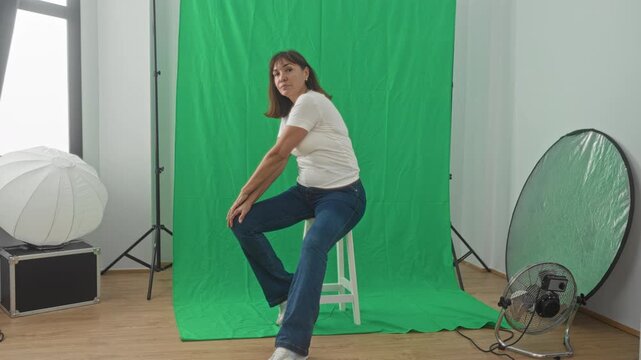 Woman seated on stool with hand on chin, blue jeans and white tee, green screen studio with reflector and fan visible; calm reflection.