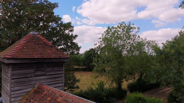 Red roof tiles stretch under blue sky with white clouds. Wooden chimney rises from thatched roof against green trees. Open field lies beyond, framed by leafy branches