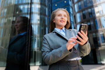 Fototapeta premium Standing confidently outside a modern building, a woman engages with her smartphone while dressed in professional attire
