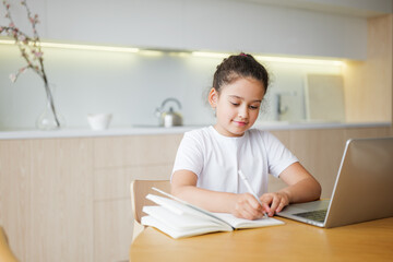 Child happily studies at a cozy kitchen table with a laptop and notebook during the afternoon © muse studio