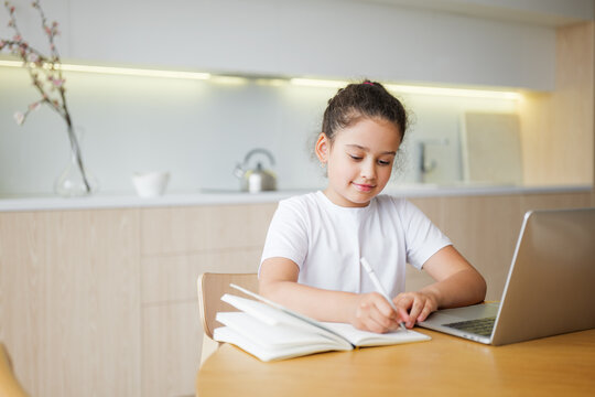 Child happily studies at a cozy kitchen table with a laptop and notebook during the afternoon