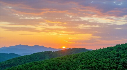 Fototapeta premium Golden sun sets behind blue mountain range over dense green forest. Vibrant orange, pink sky clouds shine during evening twilight hour. Beautiful panoramic landscape view provides calm atmosphere in
