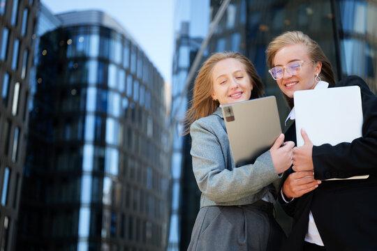 Two joyful young women sharing a moment while looking at a tablet outside modern city buildings