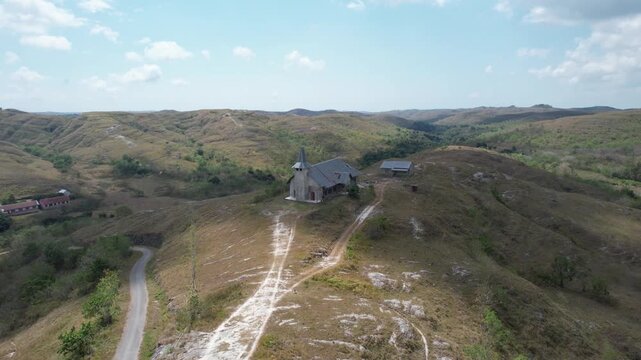 A cinematic aerial drone shot capturing the iconic Ndata Church standing gracefully atop a hill in the heart of Sumba, Indonesia. Surrounded by vast rolling savanna hills, winding rural roads, scatter
