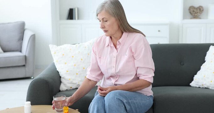 Senior woman with glass of water taking pill on sofa at home, camera moving in