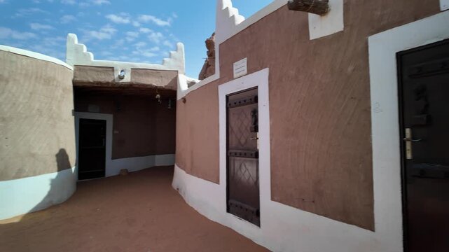 Ushaiqer Heritage Village preserving Najdi mud-brick architecture with white decorative trim, round tower and vivid red bougainvillea against a partly cloudy sky, historic charm of Saudi Arabia