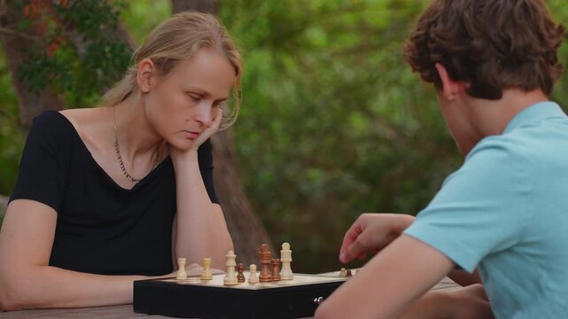 A mother and her fifteen year old teenage son play chess together at a table in a green park, focusing on their board game strategy
