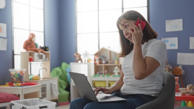 Woman holding red smartphone to ear, fingers typing on laptop while seated amid toys in a kindergarten building; focused.