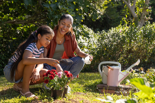 African American mom and youth daughter kneeling in yard, tending potted red blooms near water cans