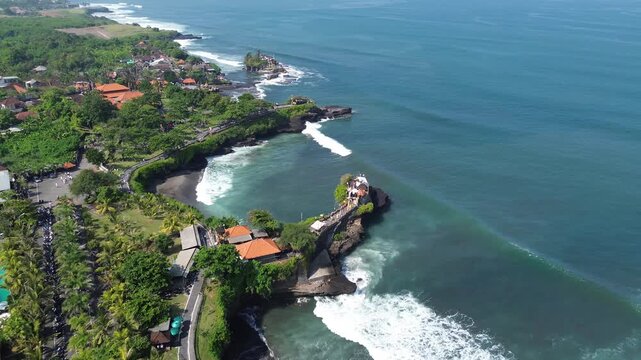 Aerial view shows crashing waves against rocky coastlines. Vibrant orange roofs contrast with lush greenery and blue waters. Cliffside structures nestle beside ocean turbulence. Tanah Lot 