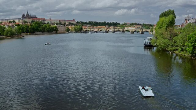 Motorboat cruising on the Vltava River with panoramic view of Prague Castle complex and historical bridges under cloudy sky.
