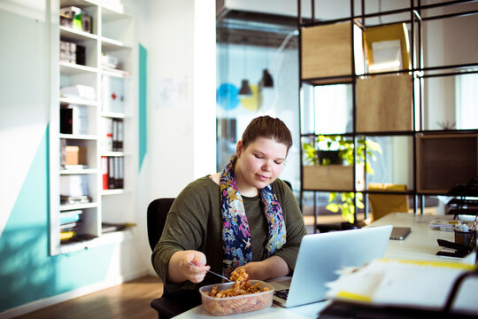 Woman eating lunch while working on laptop in modern office