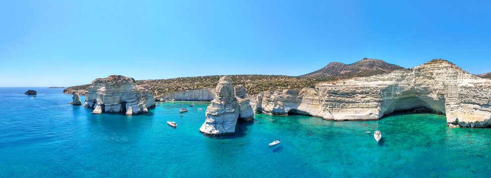 Boats float near the white limestone cliffs and caves of Kleftiko, Milos, Greece, under a clear blue sky.